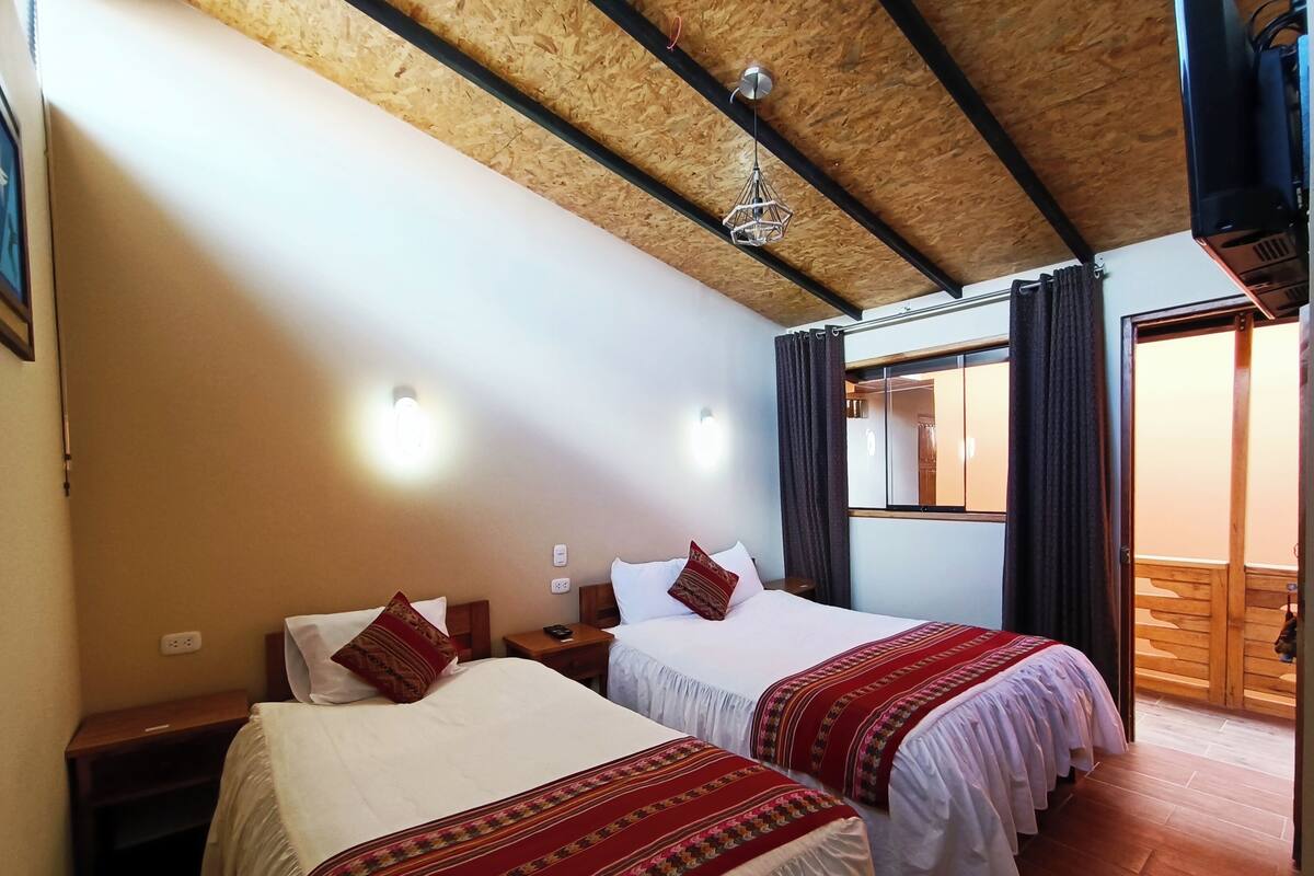 A room with two beds, wooden ceiling beams, a hanging light fixture, and a window with curtains.