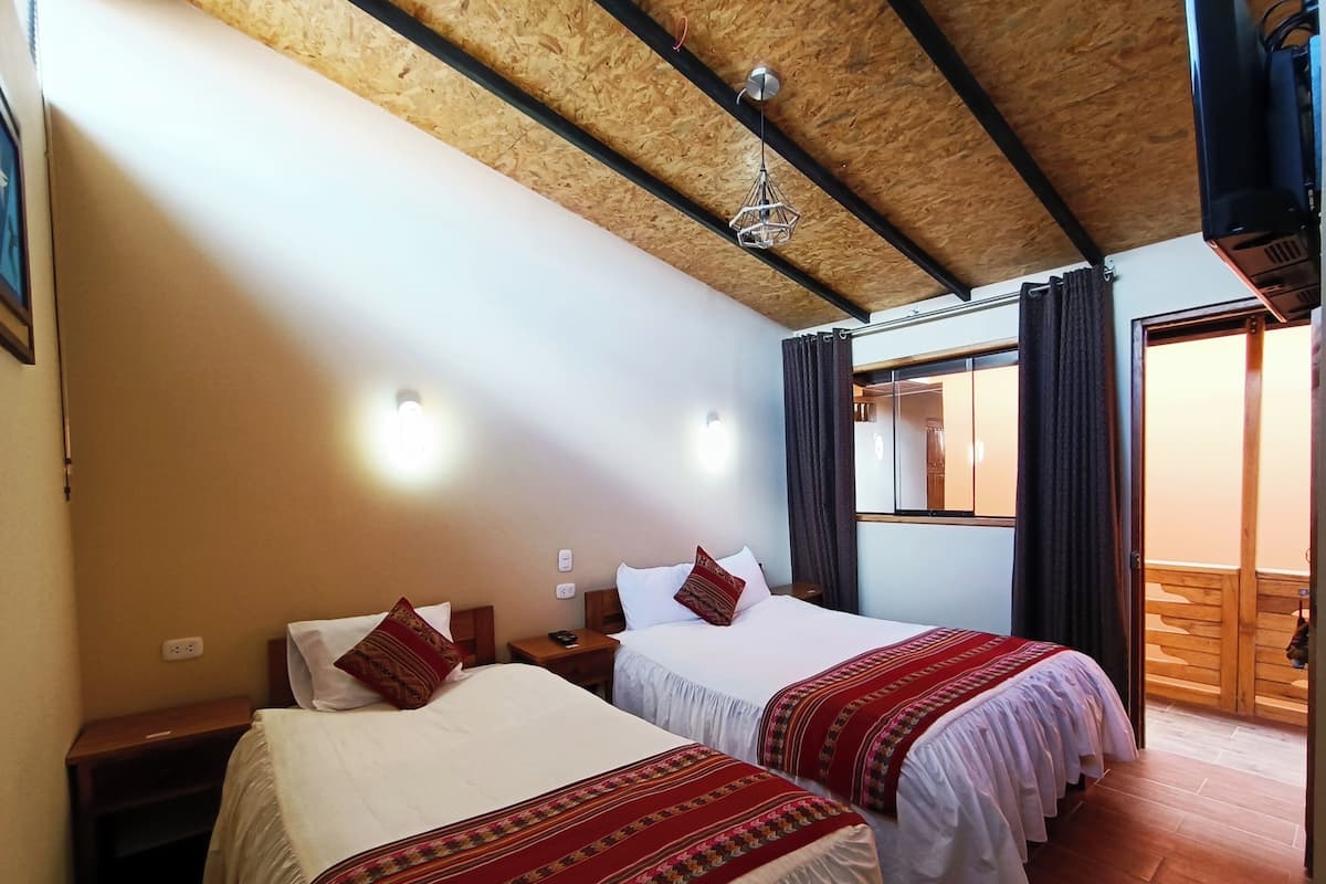A room with two beds, wooden ceiling beams, a hanging light fixture, and a window with curtains.