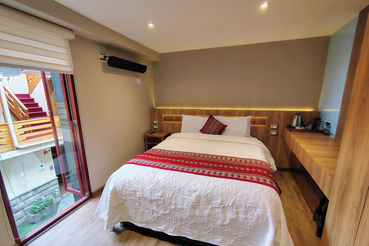 A hotel room with a bed, bedside tables, a small desk, a built-in shelf, and a sliding glass door leading to a balcony.