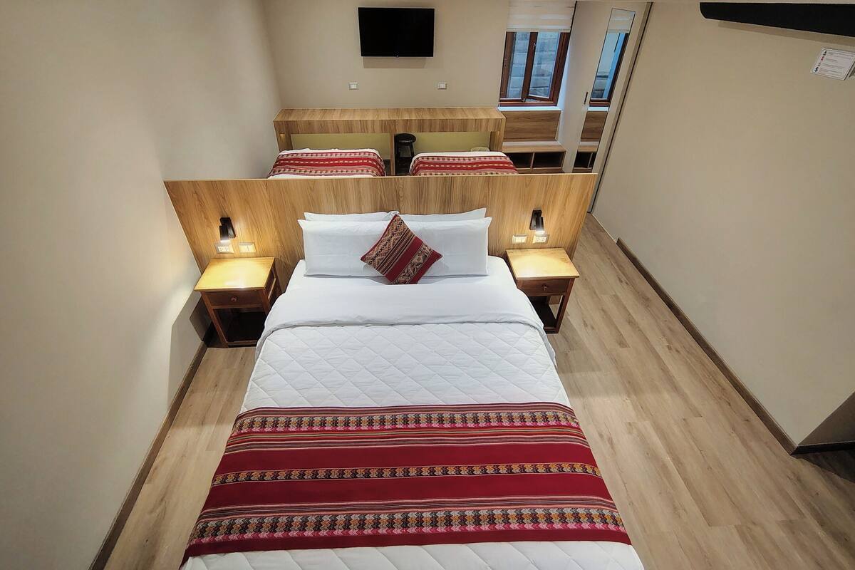 A hotel room with a large bed, two bedside tables with lamps, a flat-screen TV mounted on the wall, and a window with blinds.