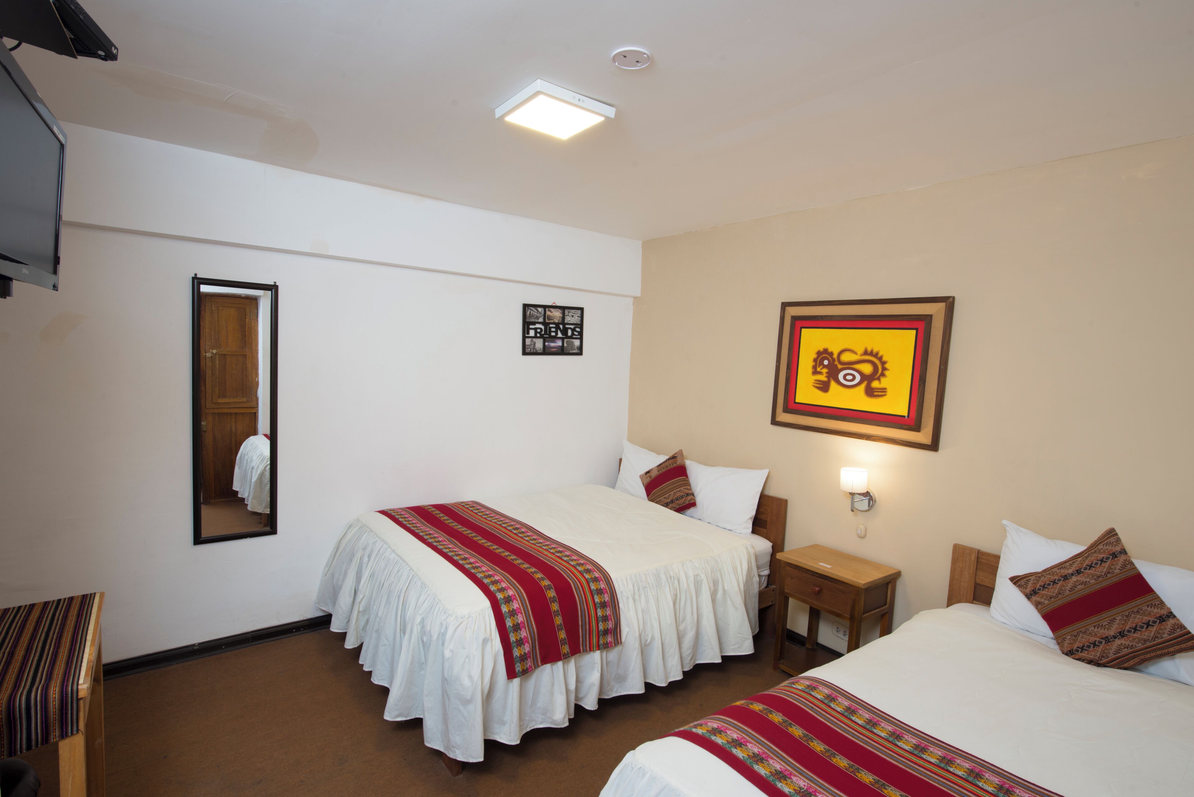 A hotel room with two beds, a bedside table, a nightstand, a mirror, a wall-mounted TV, and a framed picture.