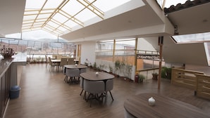 Un interior espacioso y moderno con techo alto y tragaluces, que presenta una zona central de comedor con mesas y sillas, rodeada de plantas en macetas y vistas a la ciudad.