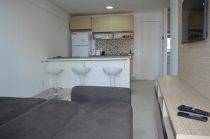 A compact kitchen with a microwave, refrigerator, and a small island with stools. There is a dining table with two remote controls on it. - Landscape Beira Mar - MeuLugarCeará (Fortaleza)