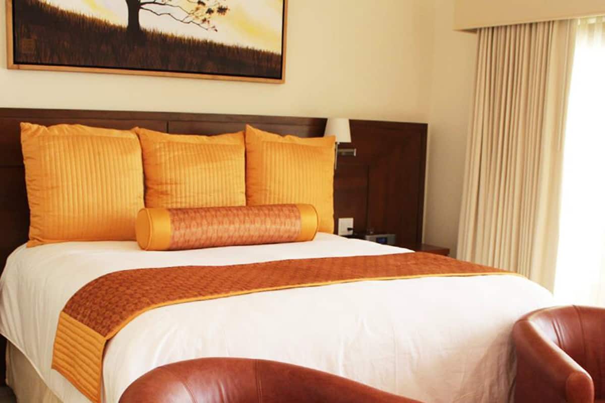 Ein Hotelzimmer mit einem großen Bett, mehreren Kissen, einem Kopfteil, einem Nachttisch, einer Lampe, einem Fenster mit Vorhängen und zwei Ledersesseln.