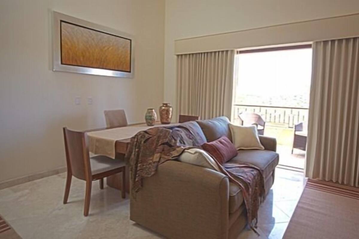 Ein Wohnzimmer mit einer Couch, einem Esstisch und Stühlen. An der Wand hängt ein Gemälde und durch große Fenster ist die Natur zu sehen.