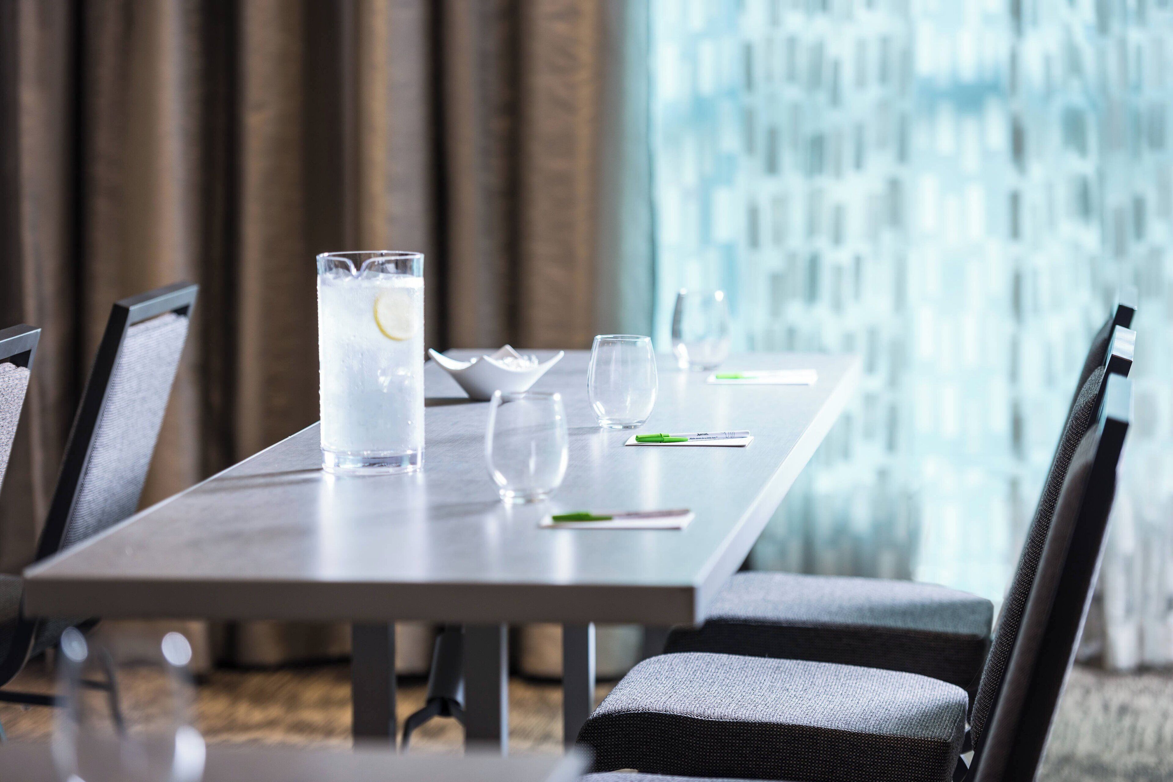 Une table à manger dressée avec une carafe d’eau, des verres et des couverts, et une vue sur un bâtiment par la fenêtre.