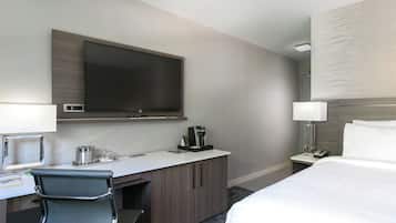 A hotel room with a bed, a desk with a chair, a television, a coffee maker, and a bedside table with a lamp.