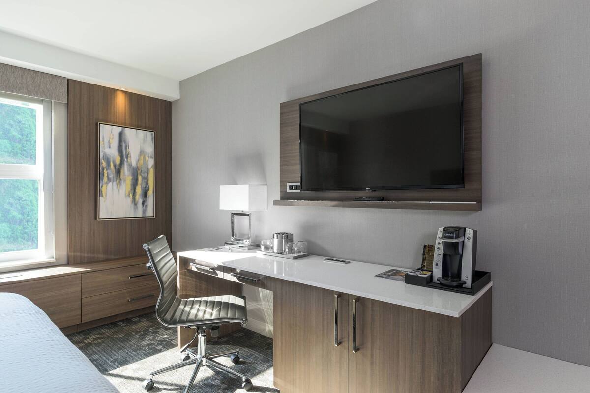 A modern hotel room with a large flat-screen TV mounted on the wall, a desk with a coffee maker, a chair, and a bed with a window offering a view of greenery.