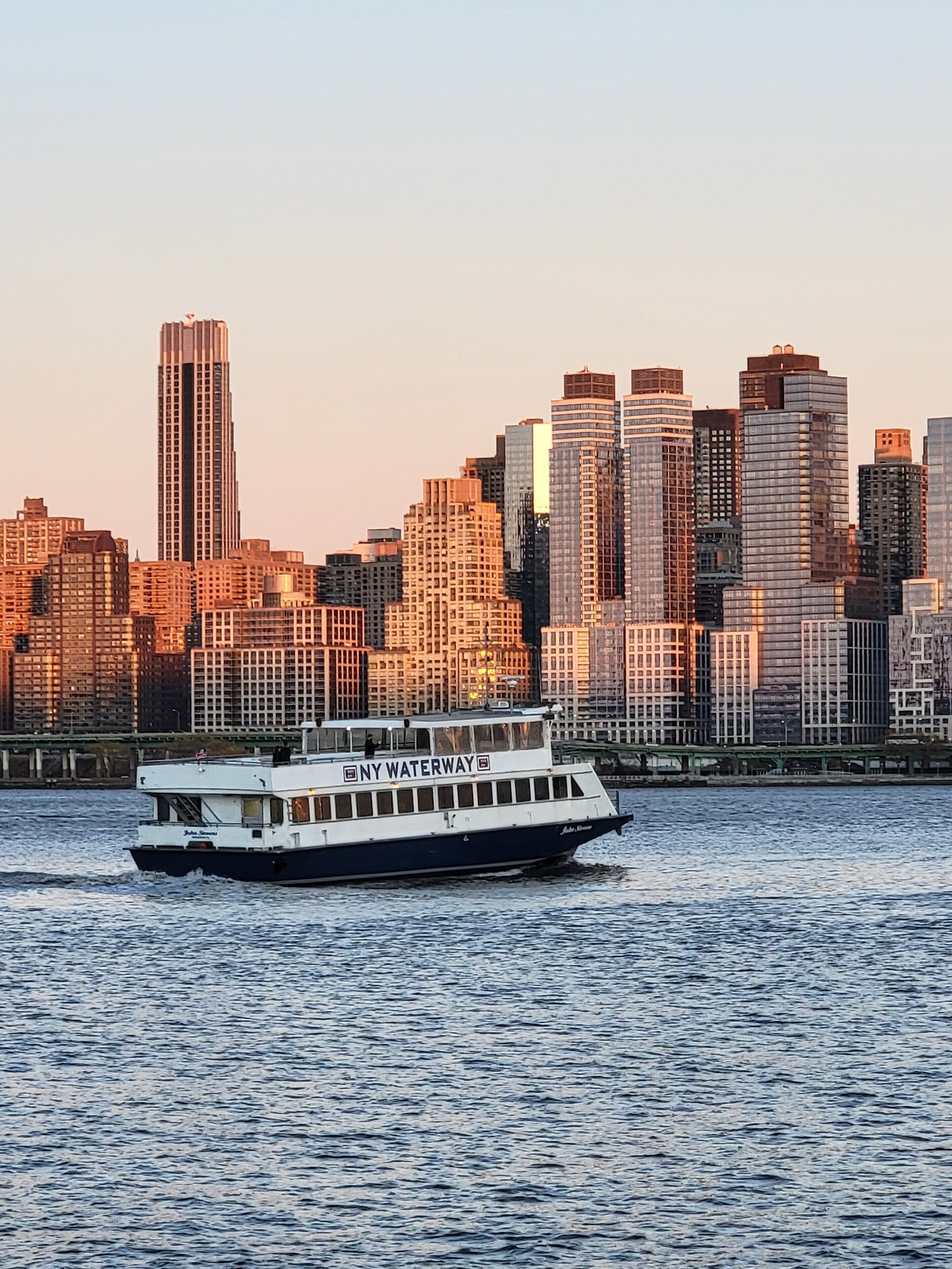 A riverboat with "NY Waterway" on its side, navigating a body of water with a city skyline in the background.