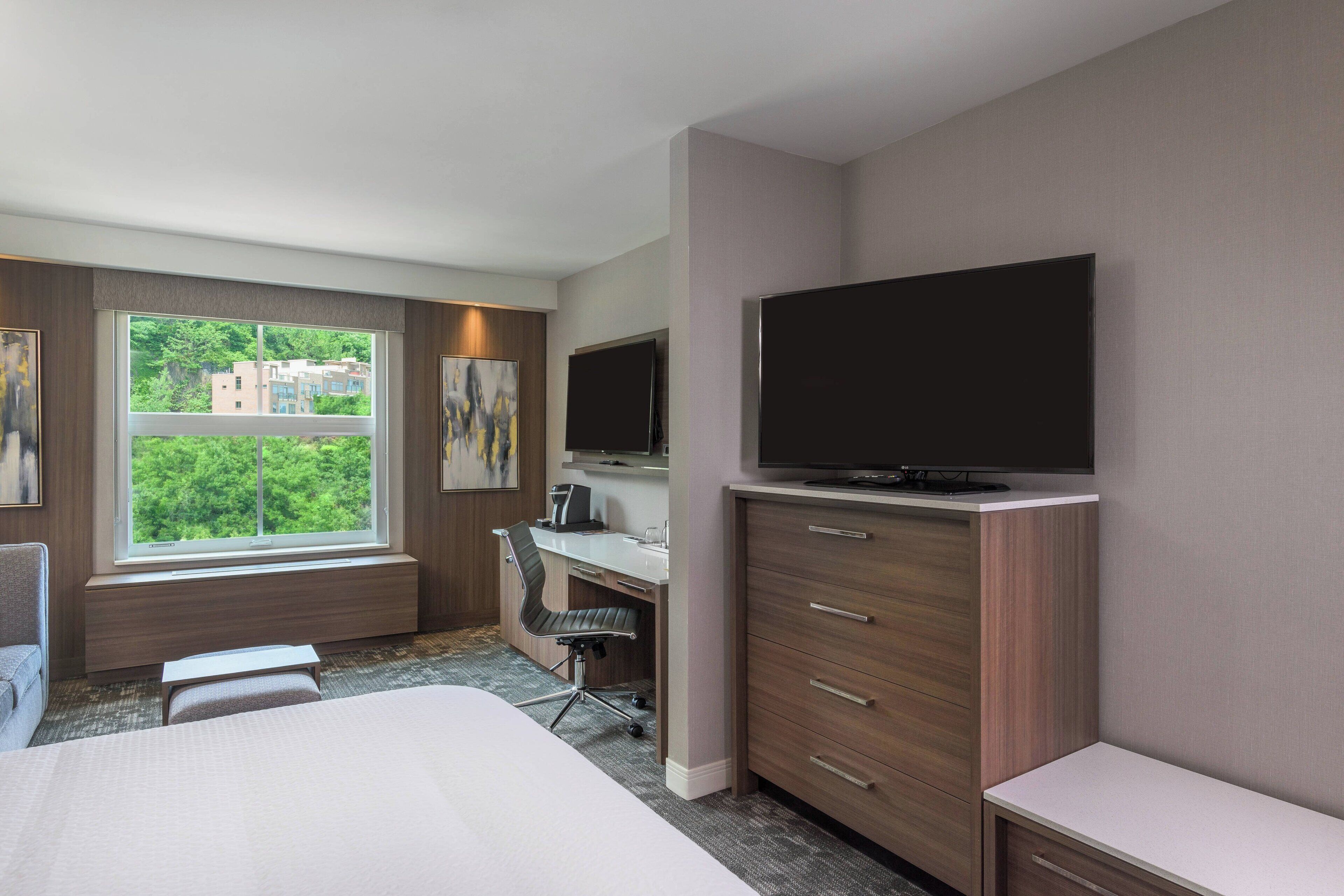 A hotel room with a bed, a desk, a chair, a television, and a window with a view of buildings.