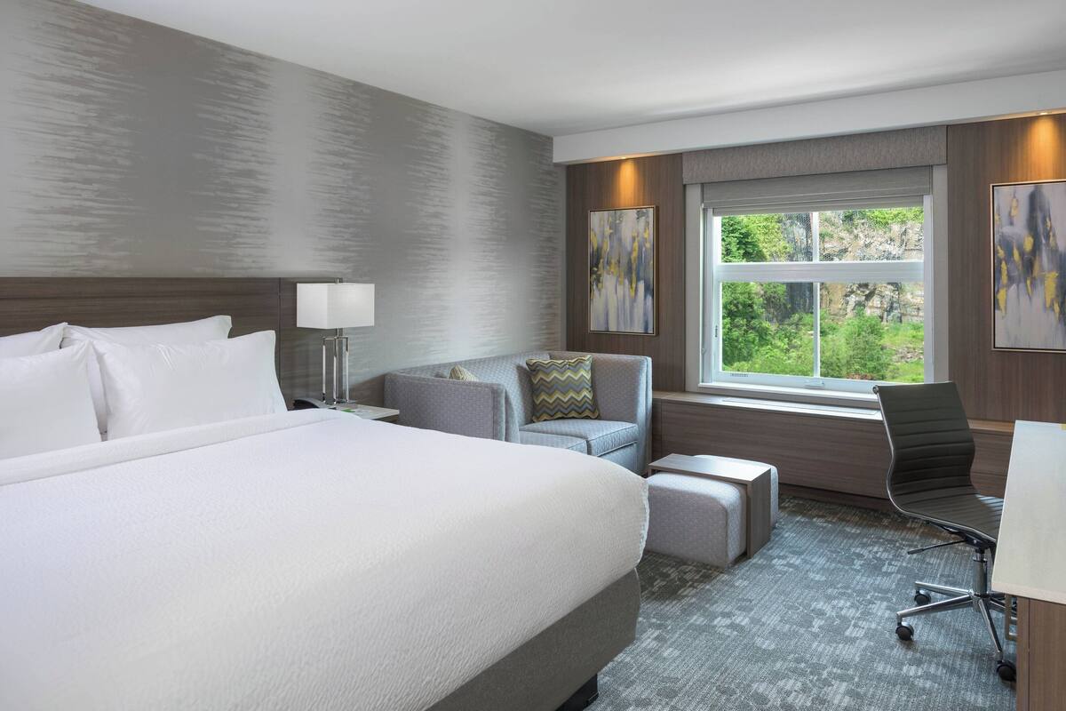 A modern hotel room with a large bed, a gray sofa, a desk, and a window with a view of greenery.