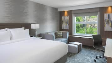 A modern hotel room with a large bed, a gray sofa, a desk, and a window with a view of greenery.