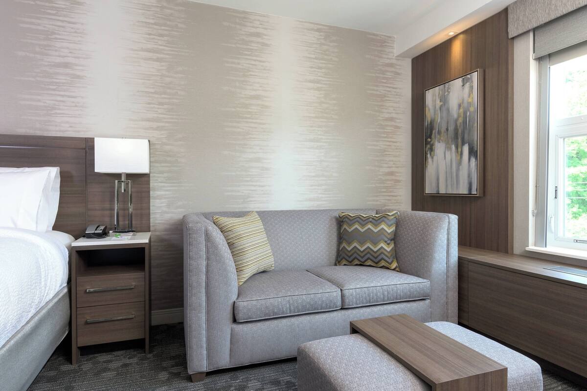 A hotel room with a bed, a grey sofa, a wooden nightstand with a lamp, a painting on the wall, and a window with a view.