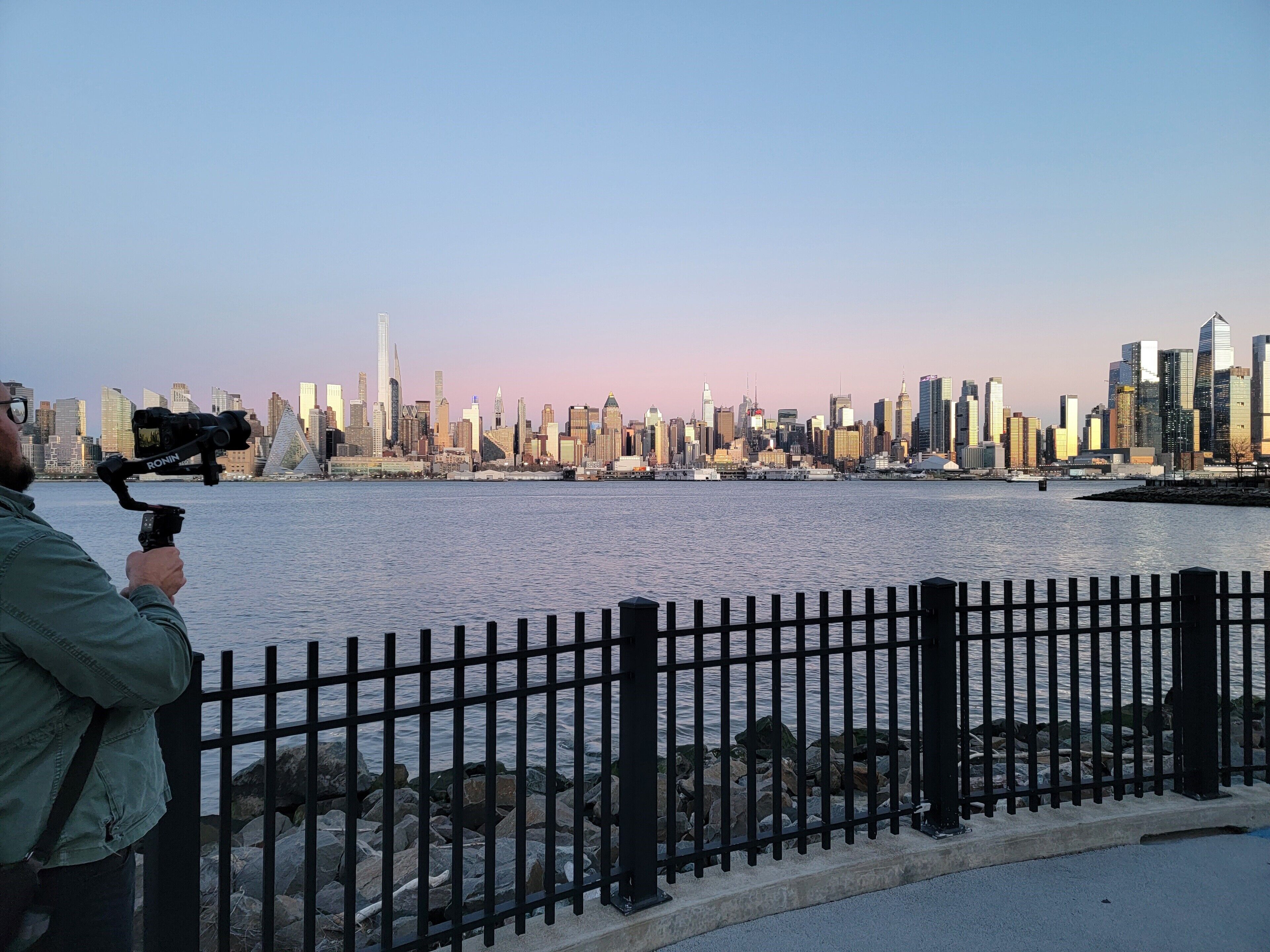 A city skyline with modern buildings across a body of water, viewed from behind a metal fence.