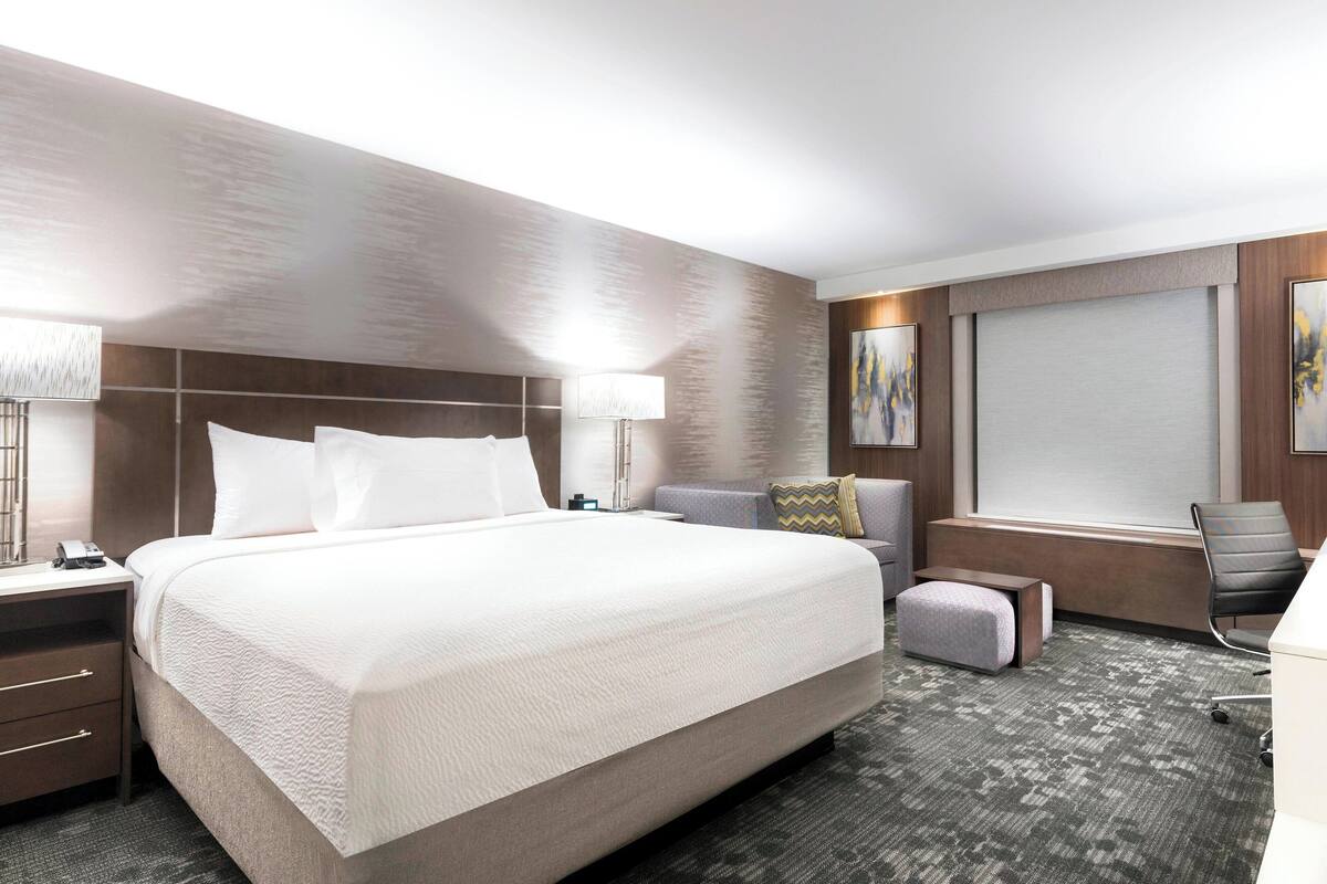 A modern hotel room with a large bed, a desk, a chair, and a window with blinds.