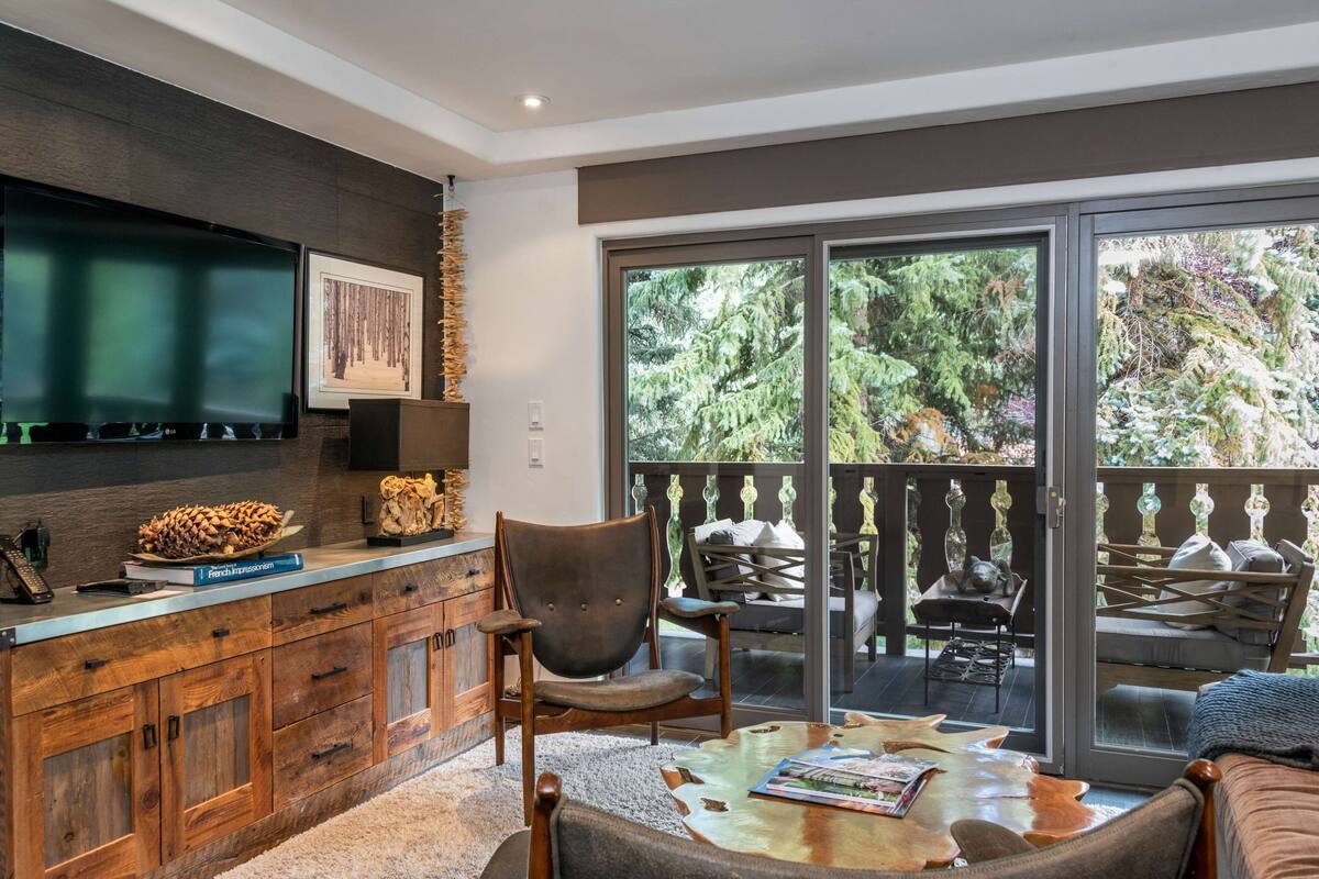 A modern living room with a large TV, wooden cabinets, a comfortable seating area, and a balcony with outdoor furniture.