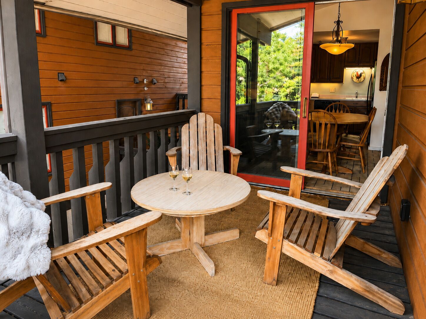 A wooden deck with a round table and four chairs, a glass door leading to a kitchen, and a hanging light fixture.