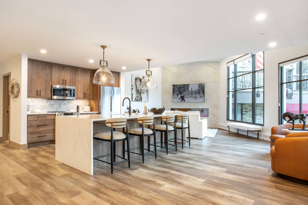 A modern kitchen with a central island, bar stools, and a seating area with a bench and orange sofa.