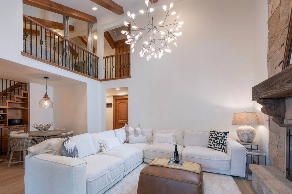 A spacious living area with a large sectional sofa, a coffee table, and a fireplace. There is a staircase leading to an upper level with a wooden railing.