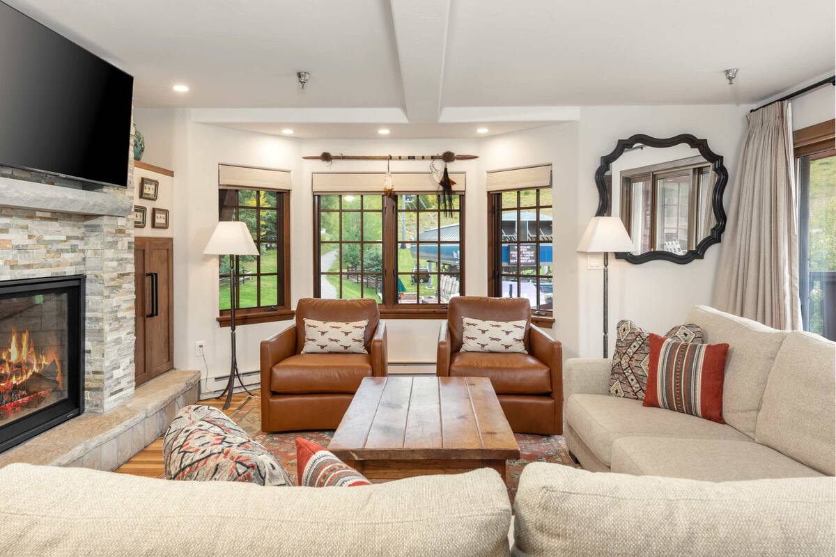 A living room with a fireplace, leather sofas, a wooden coffee table, and large windows offering a view of the outdoors.