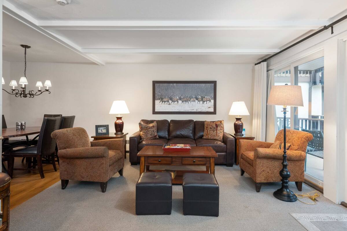 A living room with a brown leather sofa, two armchairs, a wooden coffee table, and a chandelier. There is a painting of reindeer on the wall and a window with curtains.