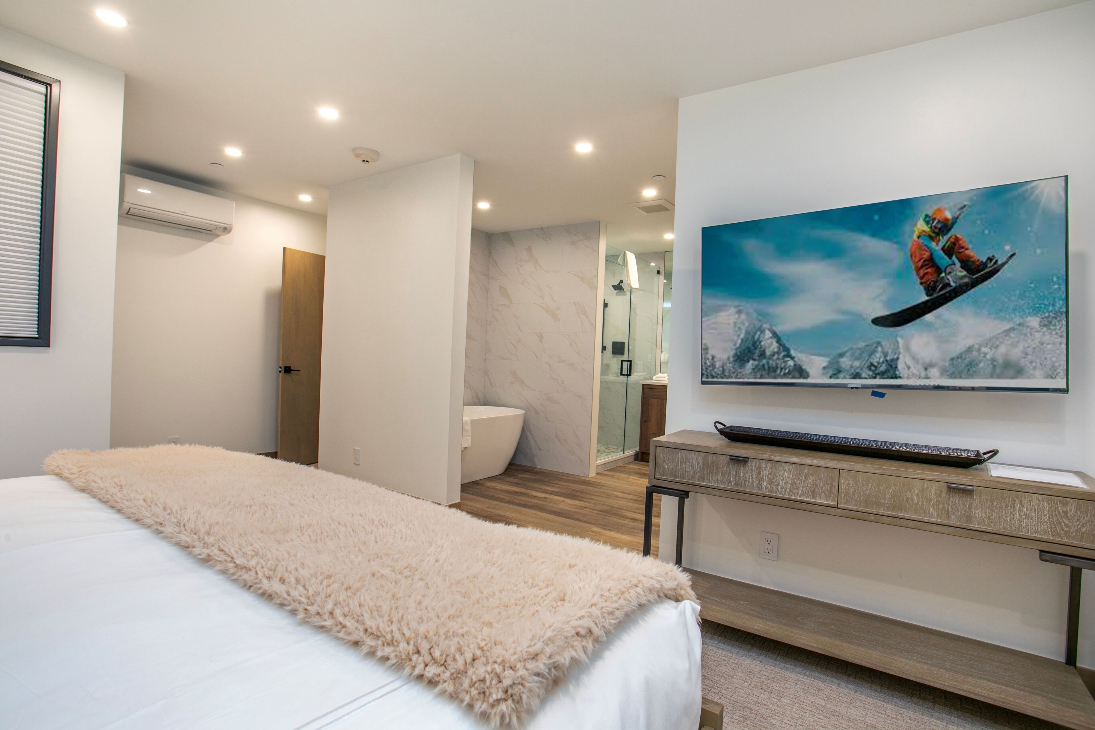 A modern hotel room with a large bed, a mounted TV displaying a snowboarder, a wooden desk, and a bathroom visible through an open door.