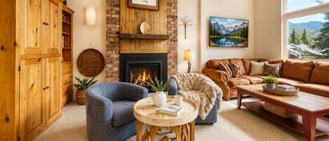 Un salon chaleureux avec une cheminée, un fauteuil bleu, un canapé marron et une vue sur les montagnes par la fenêtre.