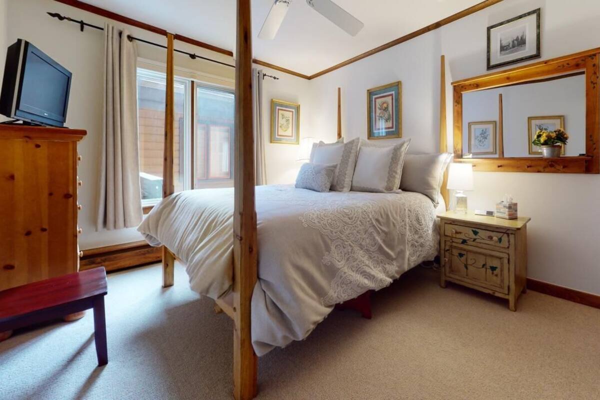 A bedroom with a large bed, wooden headboard, a bedside table with a lamp, a TV on a wooden cabinet, and a window with curtains.