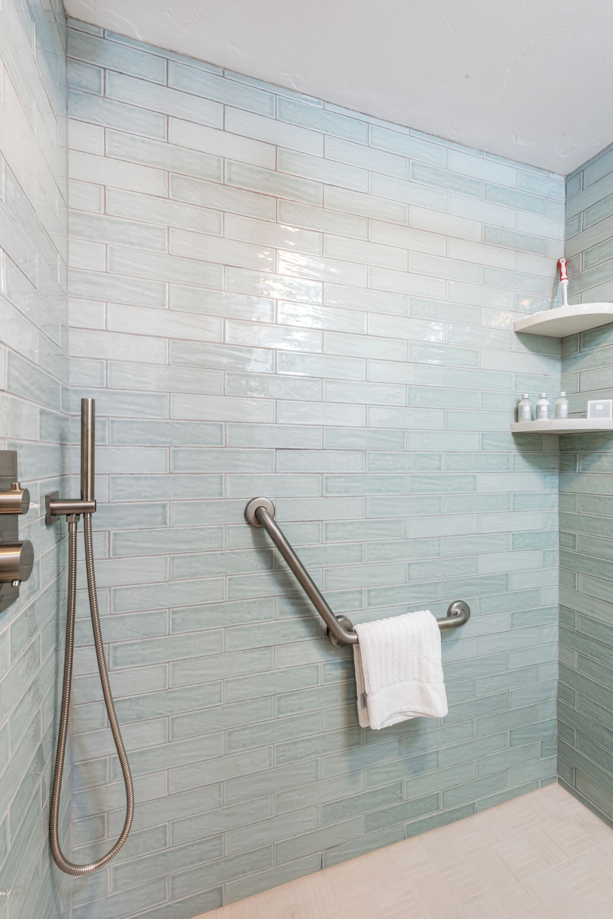 A bathroom with light blue tiled walls, a shower with a handheld showerhead, a grab bar, and a towel rack with a towel.