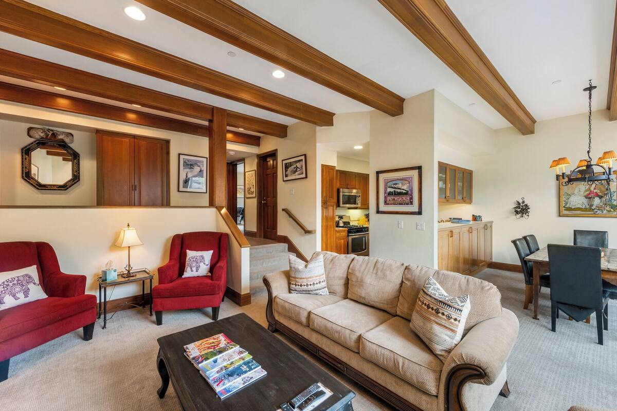 A living room with a red armchair, beige sofa, and a coffee table with magazines. There is a kitchen in the background with wooden cabinets and a dining area with a table and chairs.