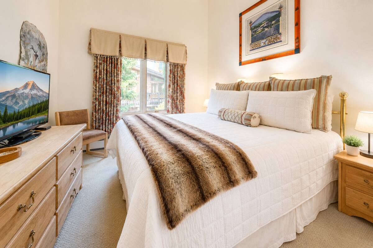 A hotel room with a bed, a desk with a TV, a window with curtains, and a framed picture on the wall.