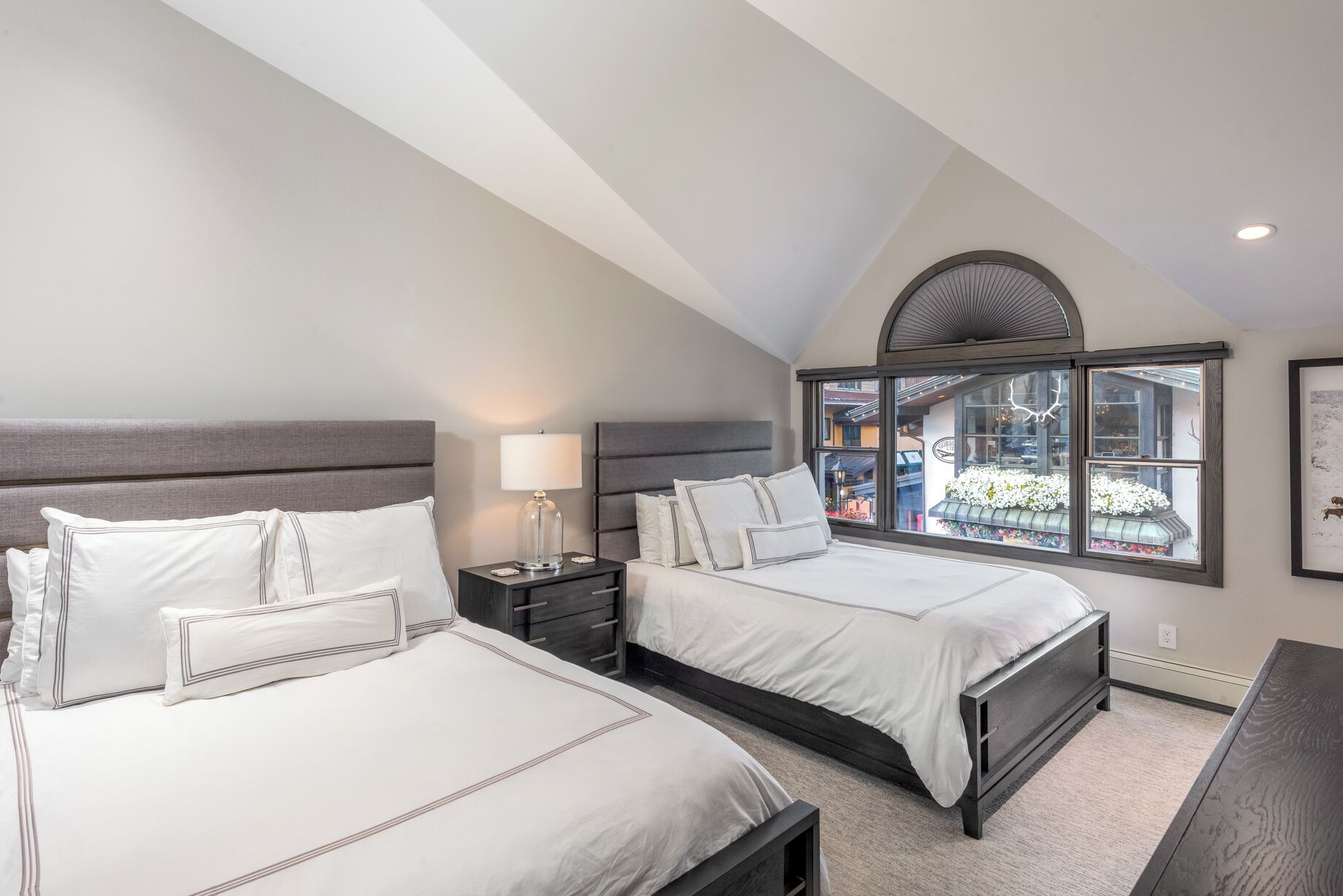 A bedroom with two beds, a nightstand, a lamp, and a large window with a view of flowers and a building outside.