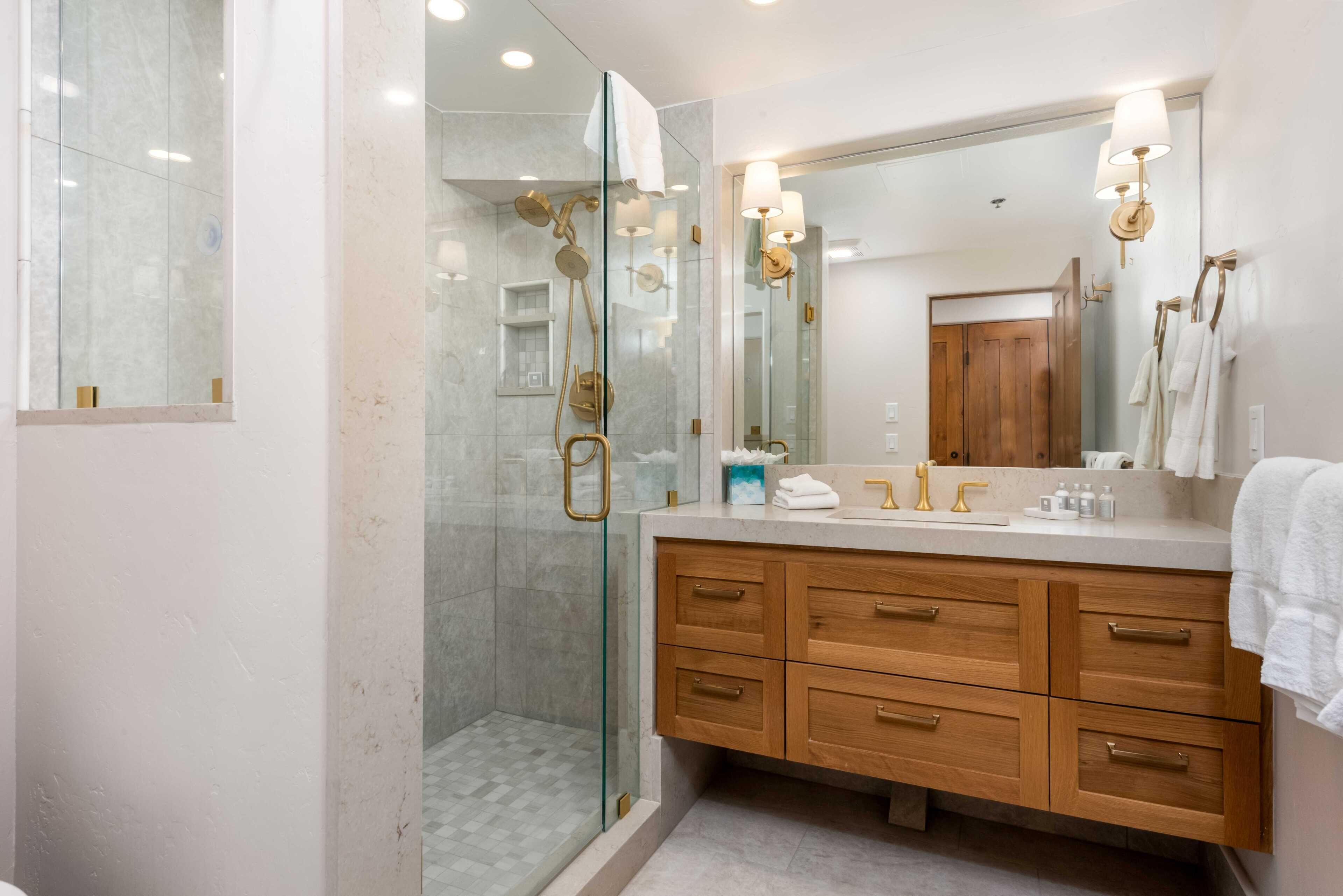 Un baño moderno con ducha de vidrio, tocador de madera con varios cajones y un espejo grande con luces empotradas.