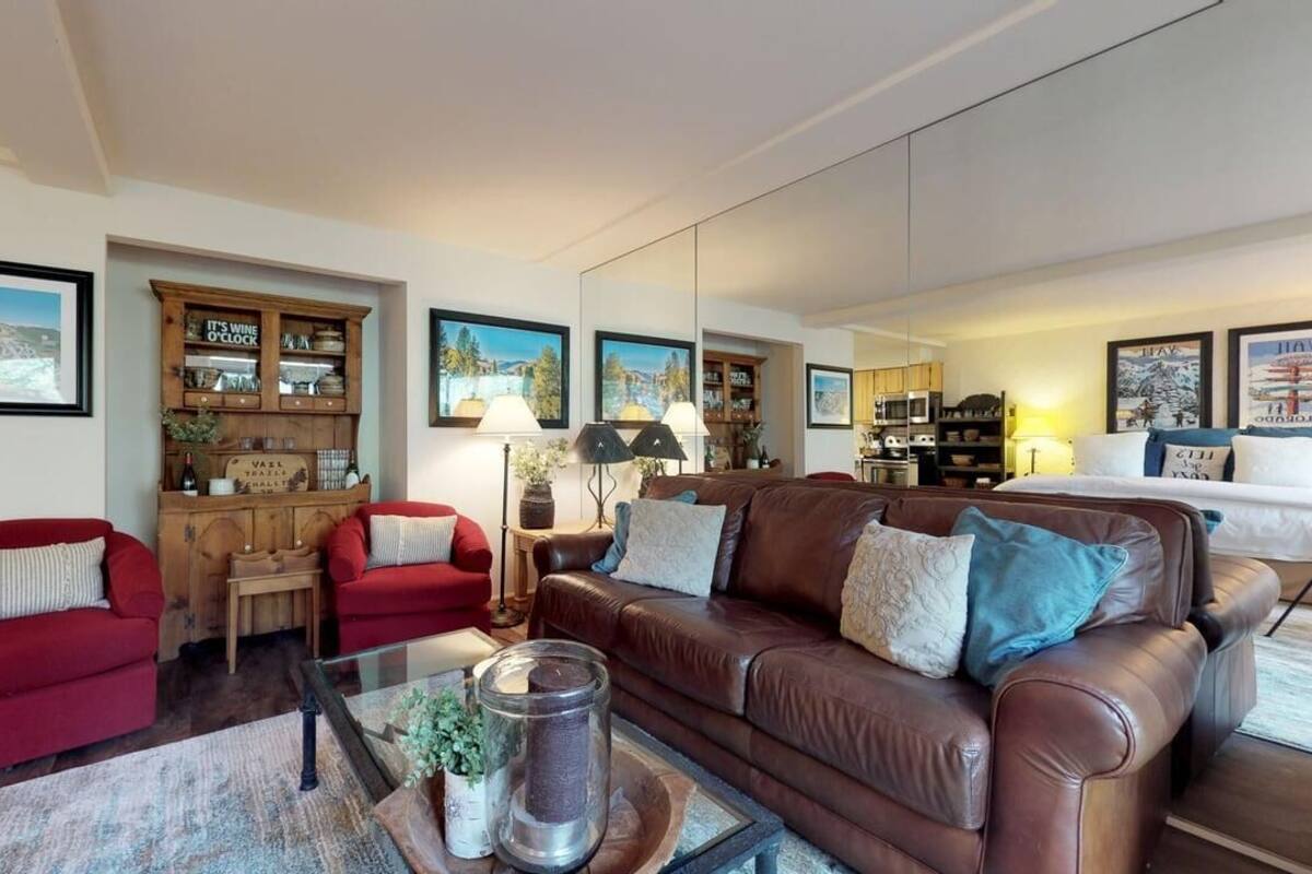A living room with a brown leather sofa, red armchair, and a glass coffee table with a candle. There are framed pictures on the walls and a wooden cabinet with glassware.