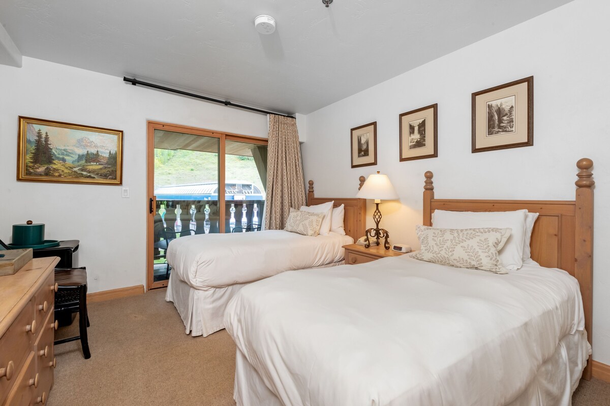 A bedroom with two beds, a dresser, a lamp, and framed pictures on the wall.