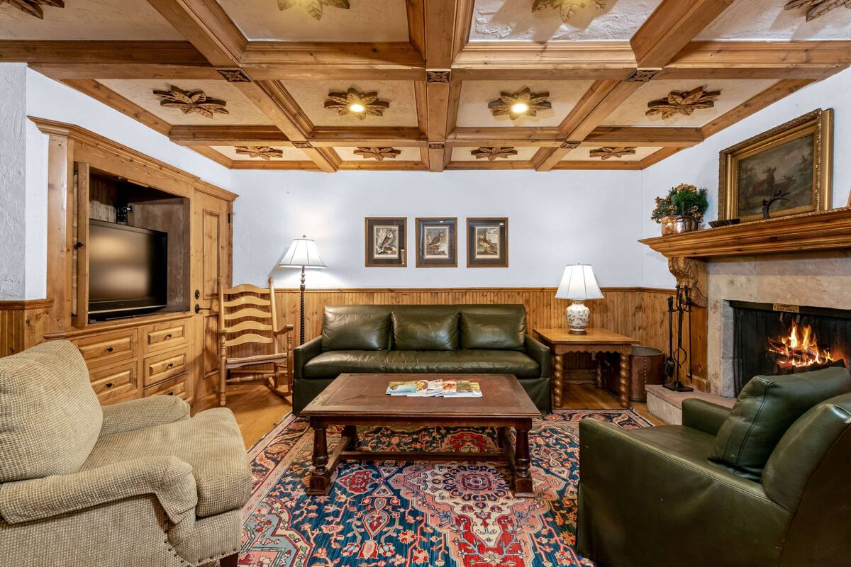 A cozy living room with a fireplace, wooden ceiling, and a patterned rug.