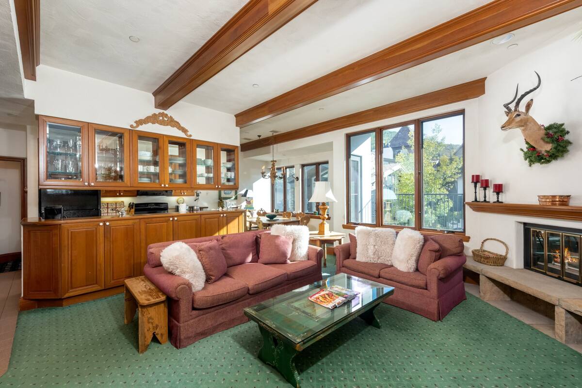 A cozy living room with two sofas, a coffee table, and a fireplace. There is a kitchen area with wooden cabinets and a television.