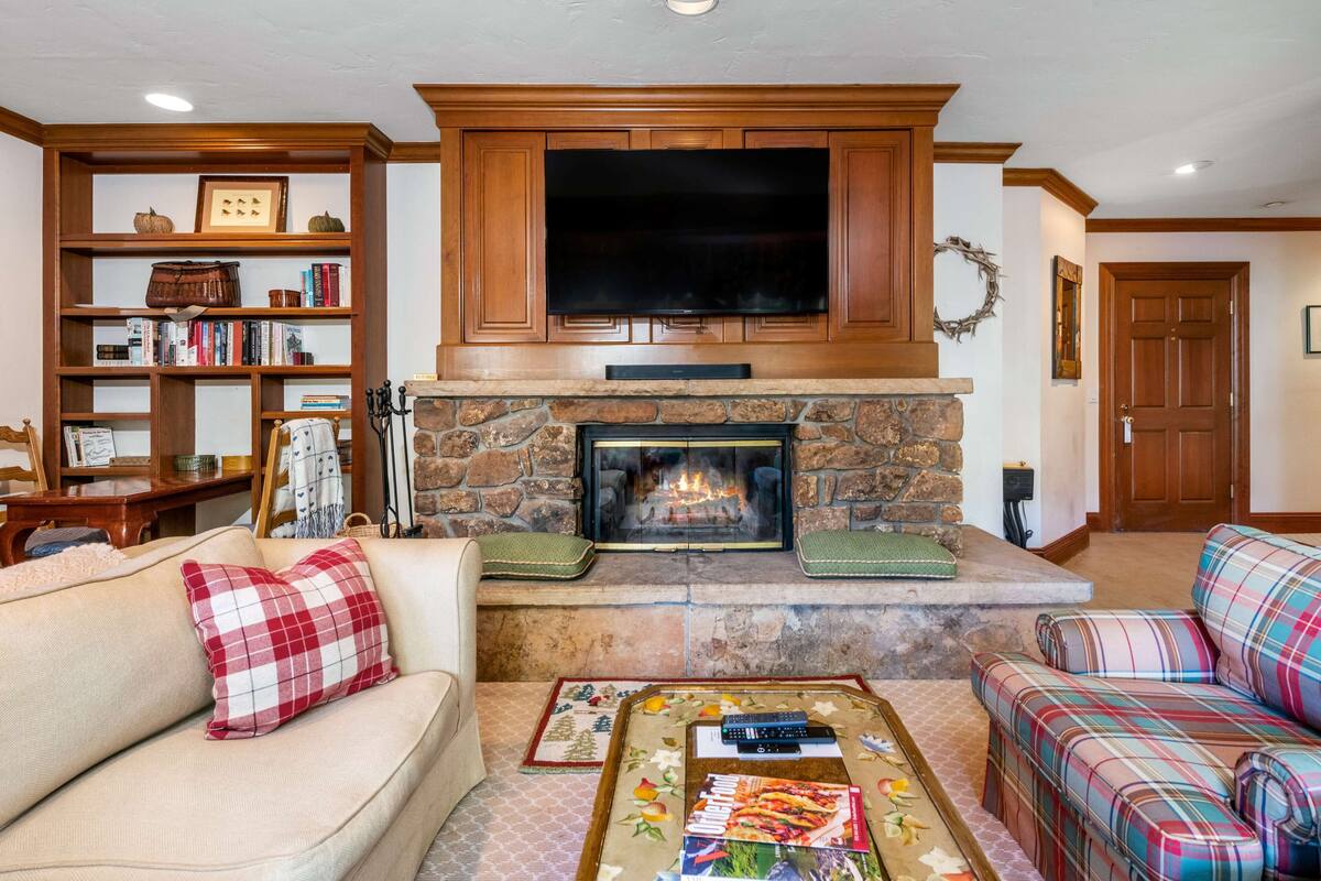 A living room with a stone fireplace, a television mounted above it, and two sofas with plaid cushions.