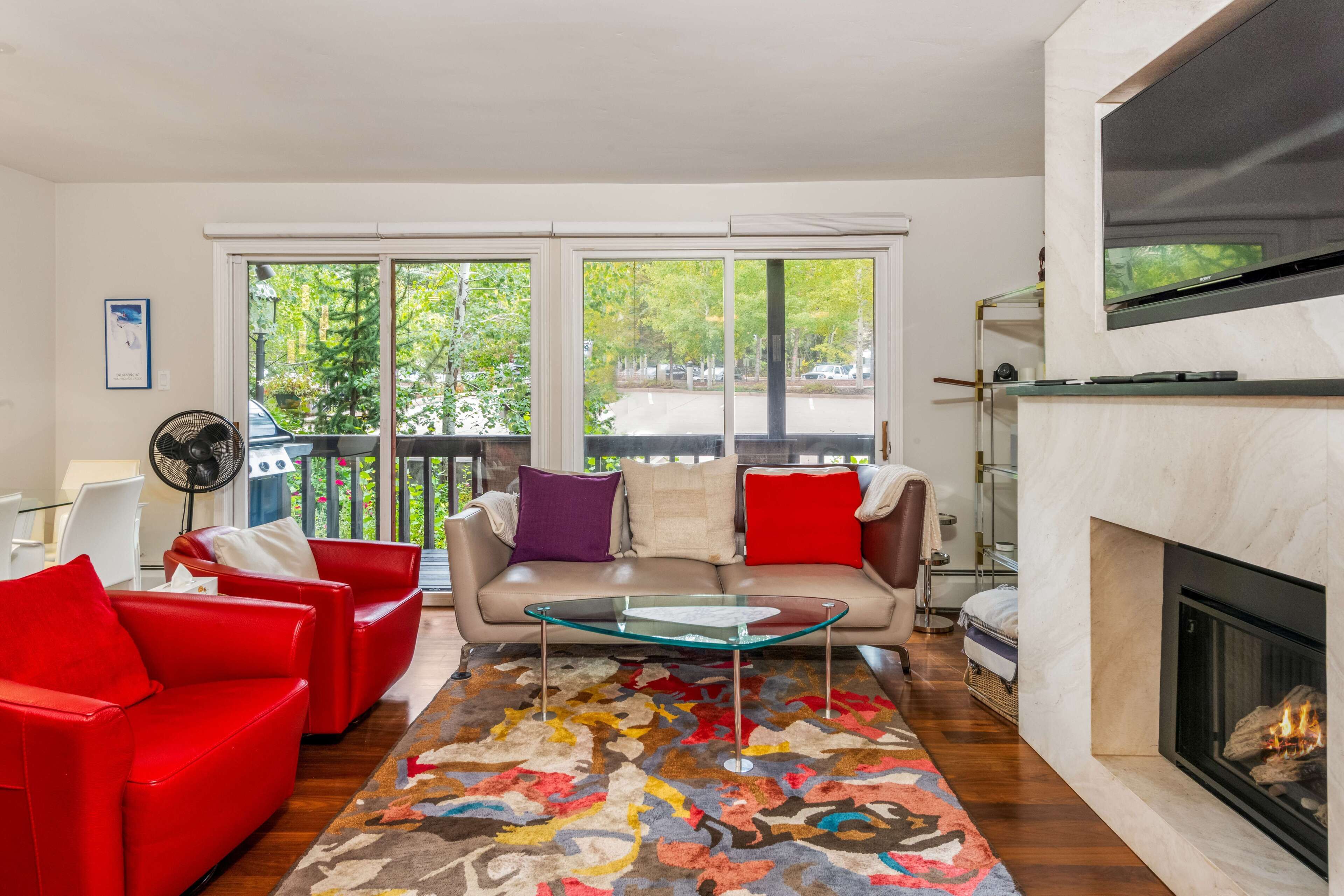 Un salon avec un canapé beige, des fauteuils rouges, une table basse en verre, un tapis coloré et une cheminée.