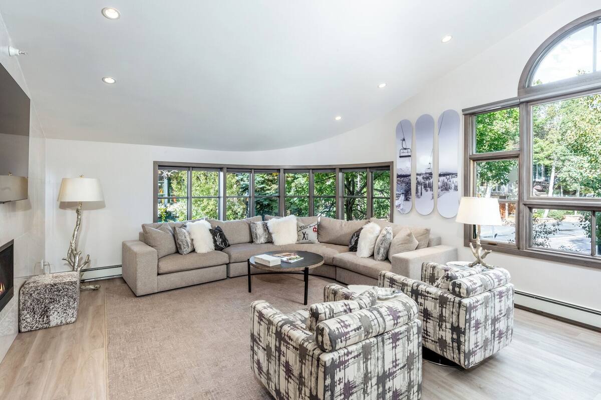 A modern living room with a large sectional sofa, two armchairs, a coffee table, and a fireplace. Large windows provide natural light and views of greenery outside.