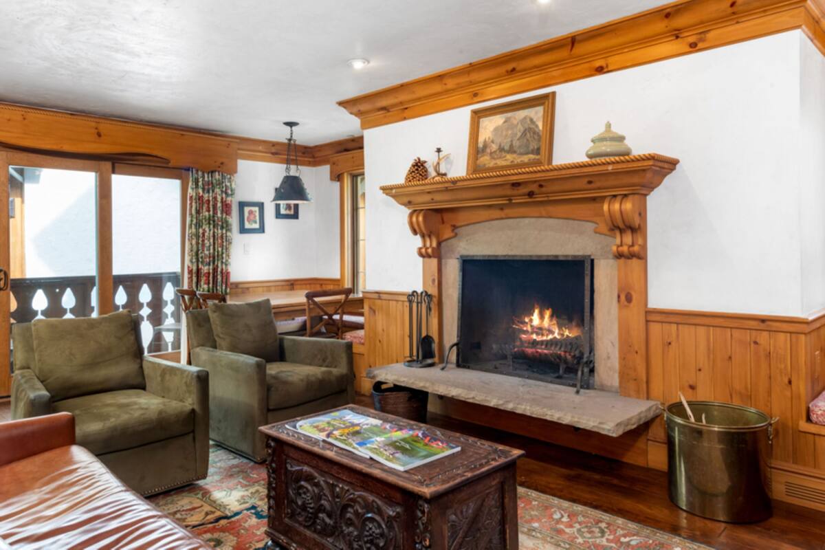 A cozy living room with a fireplace, wooden furniture, and a view through large windows.