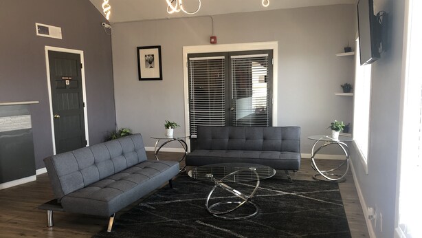 A modern living room with a grey sofa, a black rug, and a glass coffee table. There are two potted plants, a framed picture on the wall, and a window with blinds. - Extend a Suites & RV Park (Albuquerque)