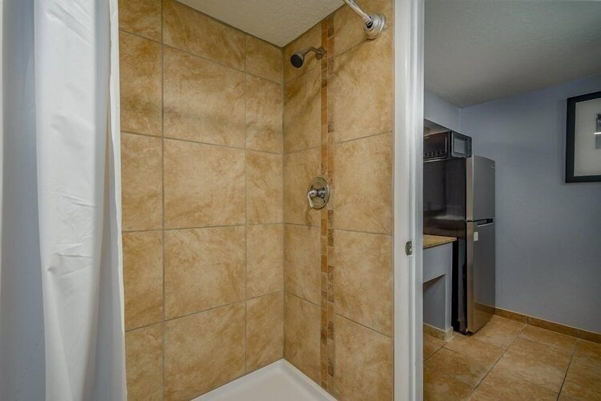 A bathroom with beige tiles, a shower, and a built-in kitchen area with a refrigerator and microwave.