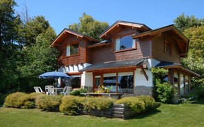 Une maison en bois à deux étages, dotée d’un balcon, d’une terrasse avec des chaises et d’un jardin.