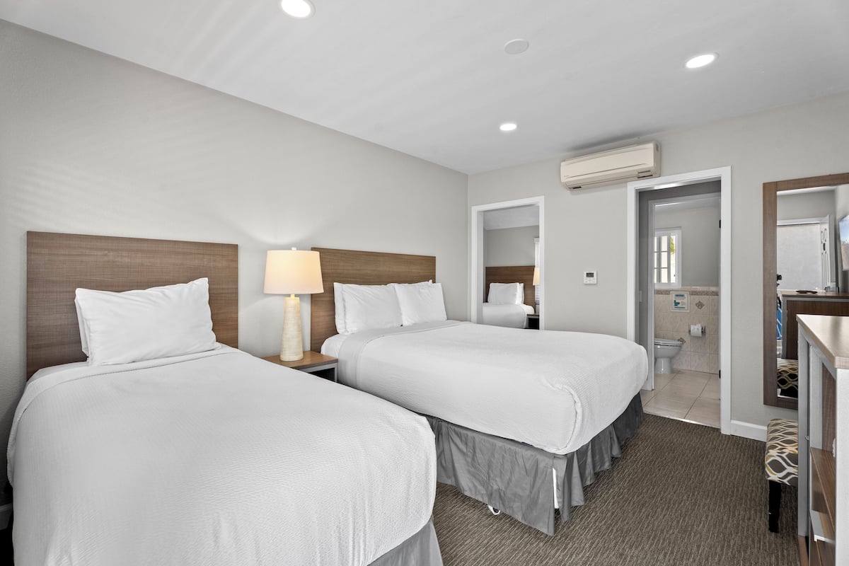A hotel room with two beds, a bedside table with a lamp, a bathroom with a toilet and sink, and a mirror.