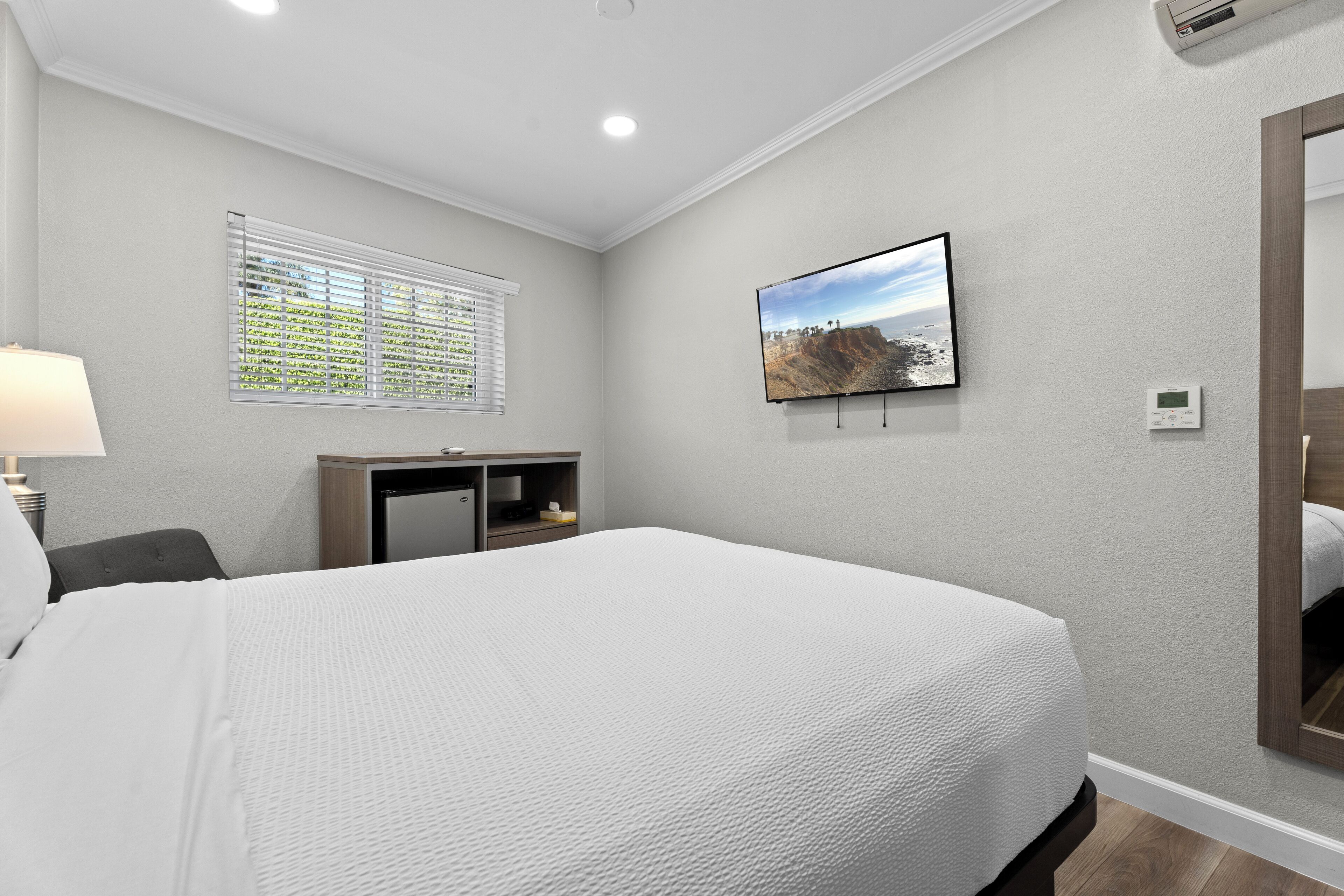 A hotel room with a large bed, a desk with a chair, a TV mounted on the wall, and a window with blinds.