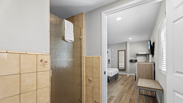 Un baño moderno con ducha, inodoro y lavamanos. En la habitación contigua hay una cama.
