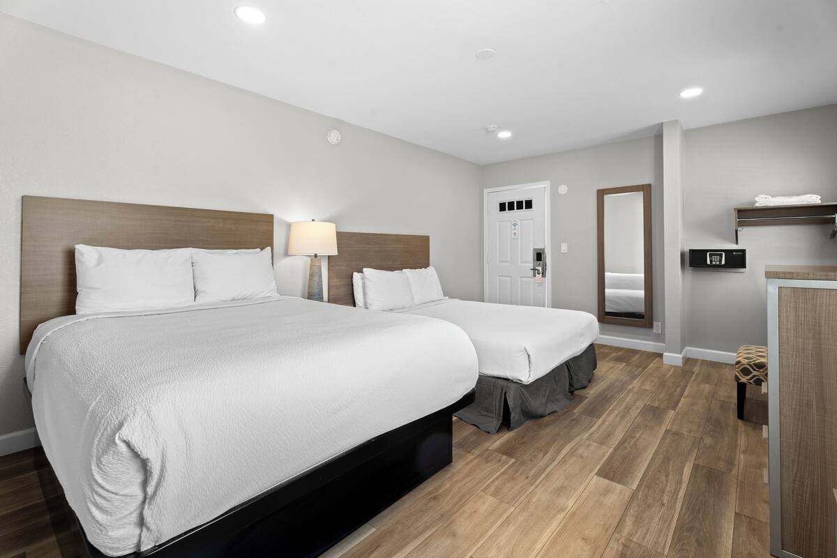 A hotel room with two beds, a wooden headboard, a bedside table with a lamp, a wardrobe, and a mirror.
