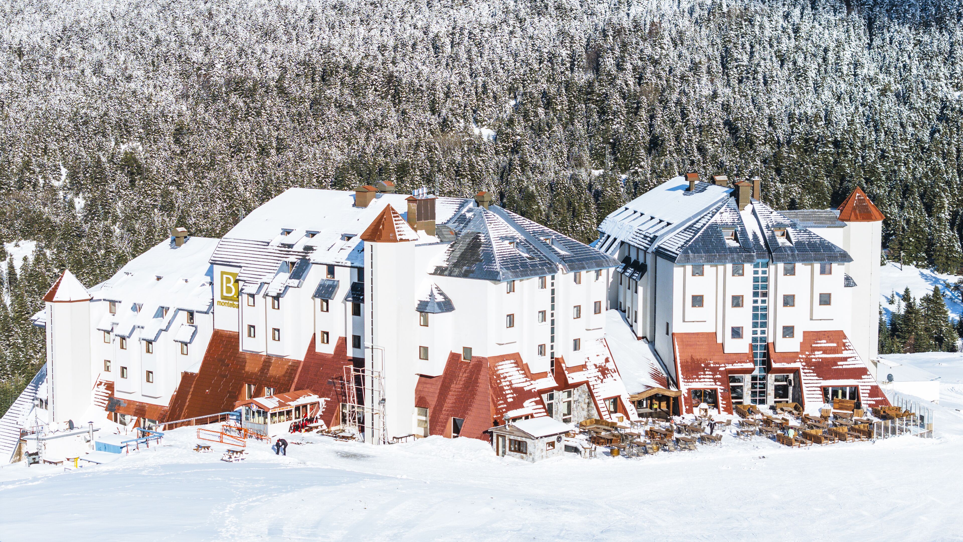A snow-covered building with multiple levels and a mix of red and white roofs, surrounded by a snowy forest.