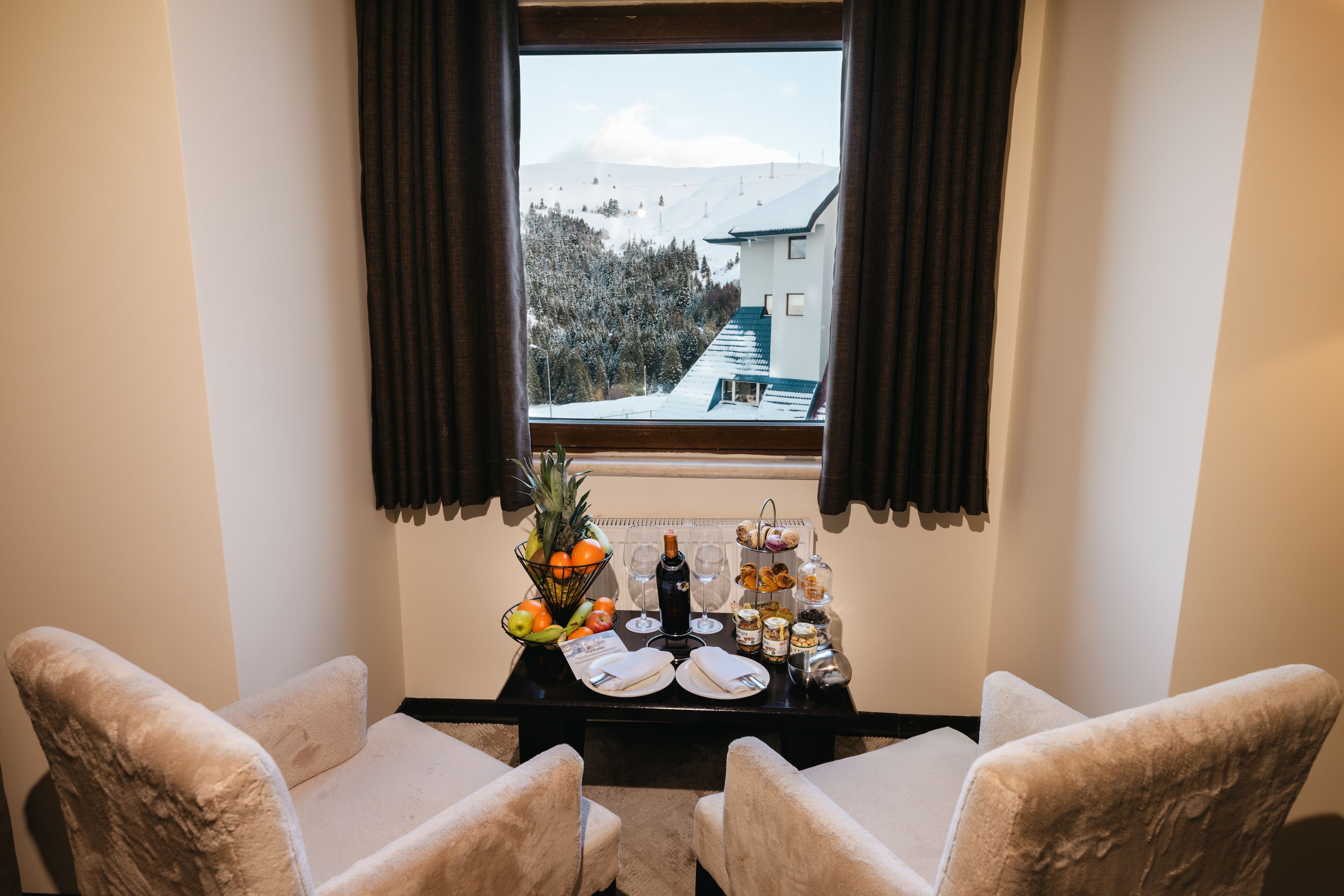 A dining area with two chairs, a table with a fruit basket, wine, and pastries. A window reveals a snowy mountain and a building.